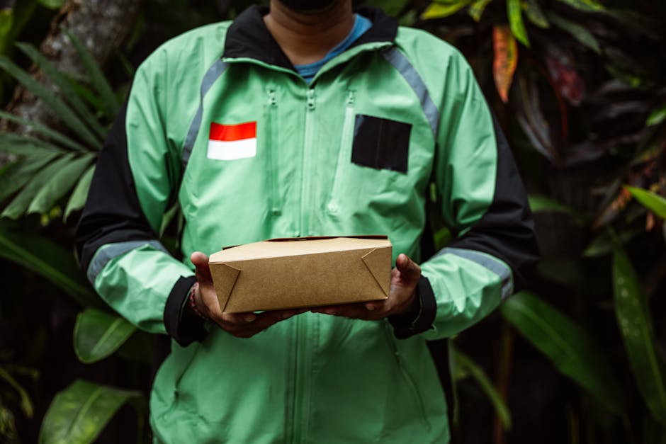 A delivery courier wearing a bright green jacket with black accents and a red and white flag patch on the chest is holding a rectangular cardboard box with both hands. The background features lush gre