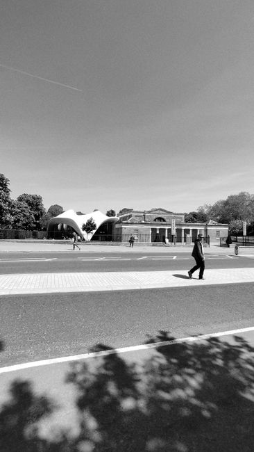 A black and white photograph depicting a street scene with several pedestrians walking along the pavement. In the background, there is a modern building with a curved, tent-like roof structure surroun