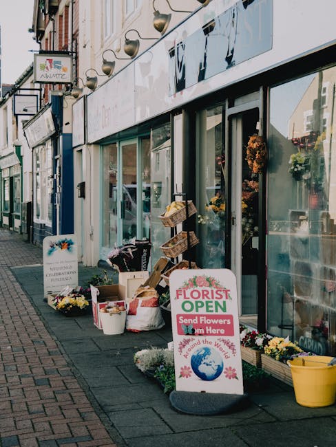 The exterior of Raynes Park Florist features a glass-fronted shop with a variety of fresh flower arrangements visible through the windows. Outside, there are multiple bouquets and floral displays in w