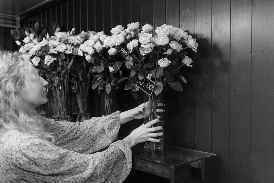 A woman with curly hair in a patterned long-sleeve blouse arranges a tall glass vase filled with a large bouquet of fresh white roses and greenery, situated on a dark wooden surface against a wooden-p