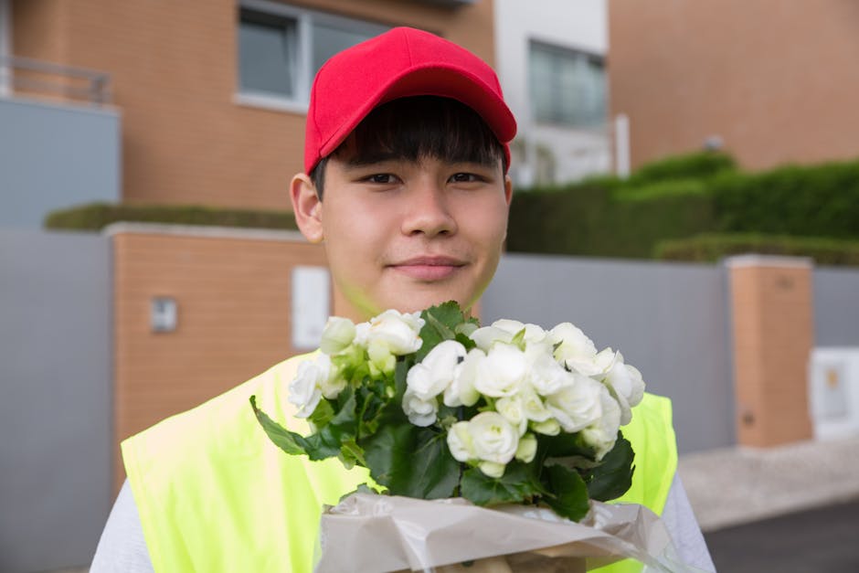 A young male flower delivery courier wearing a red cap and high-visibility yellow vest is holding a bouquet of fresh white roses with green leaves, wrapped in white paper. He is standing outdoors in a
