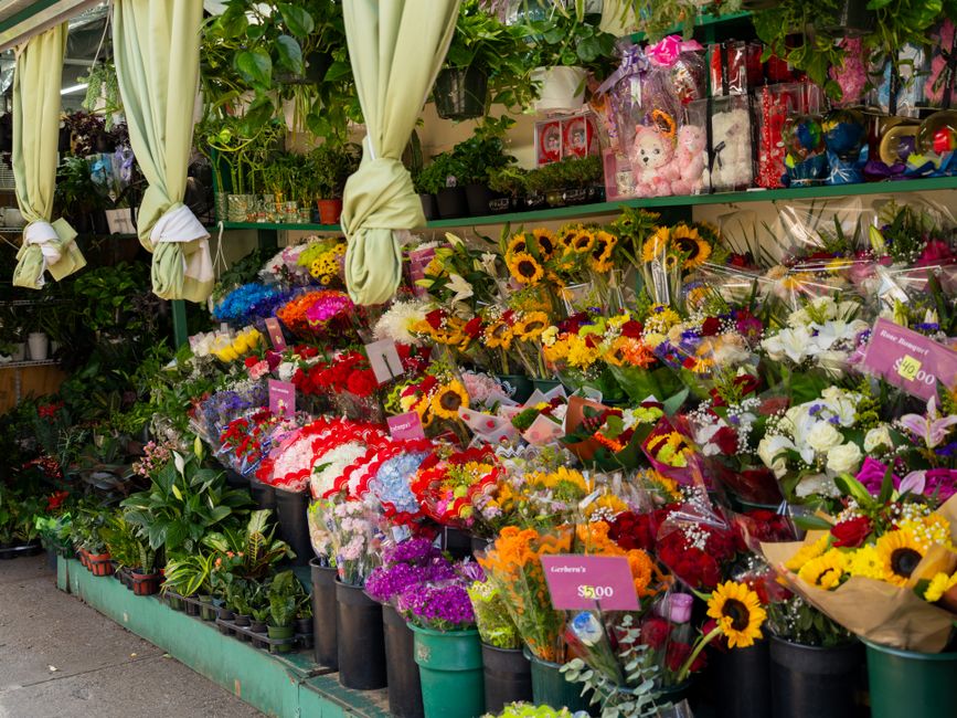 A vibrant outdoor flower market stall displaying a diverse array of fresh flowers including bright yellow sunflowers, red roses, white lilies, purple statice, and multicolored mixed bouquets wrapped i