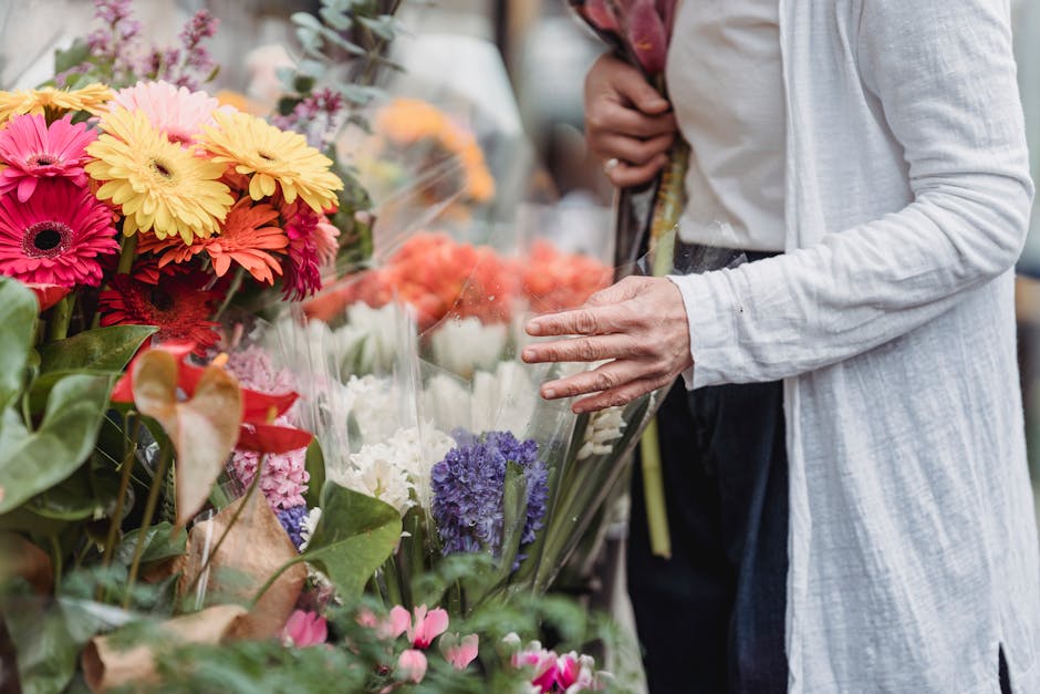 A person dressed in a light grey cardigan and dark trousers is browsing a vibrant floral display at a flower market. The arrangement includes bright yellow, pink, and coral gerbera daisies, white and