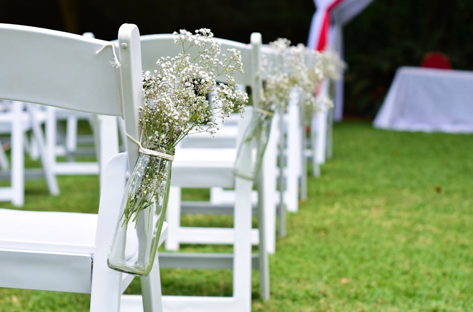 A row of white wooden chairs at an outdoor wedding ceremony venue, each decorated with a small glass vase containing delicate white baby's breath flowers, attached to the side with twine. The setting
