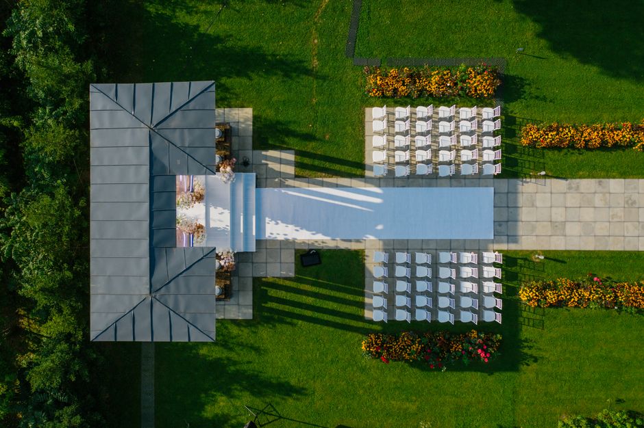 Aerial view of an outdoor wedding ceremony setup with a floral aisle leading to a decorated altar under a grey pavilion. Surrounding the arrangement are rows of white chairs, neatly aligned on a stone